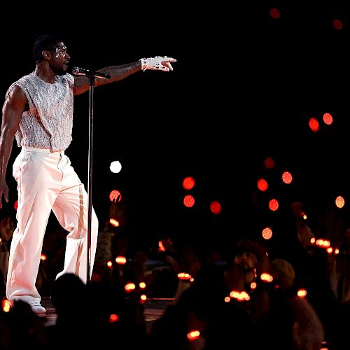 A performer in white attire sings into a microphone on stage, surrounded by an audience holding illuminated candles.