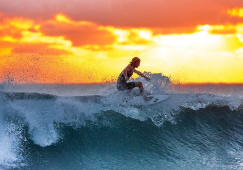 A surfer rides a wave in the ocean at sunset, with the sky glowing orange and yellow behind them.