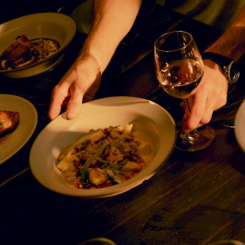 A person&rsquo;s hand reaching for a plate of food beside a glass of wine at a dim, candlelit dinner.