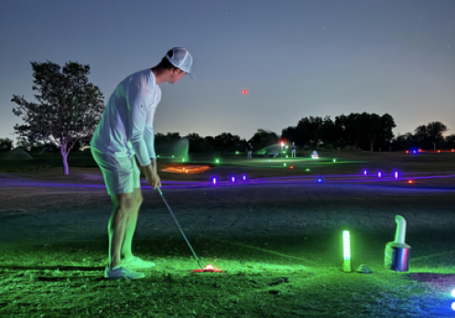 A golfer swings on a night course under colorful glow lights and glowing markers, illuminated greens and a dark sky.
