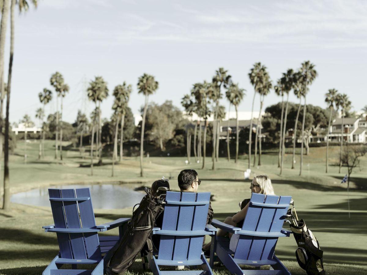 Three people sit on blue chairs near a golf course, with golf bags beside them and palm trees in the background.