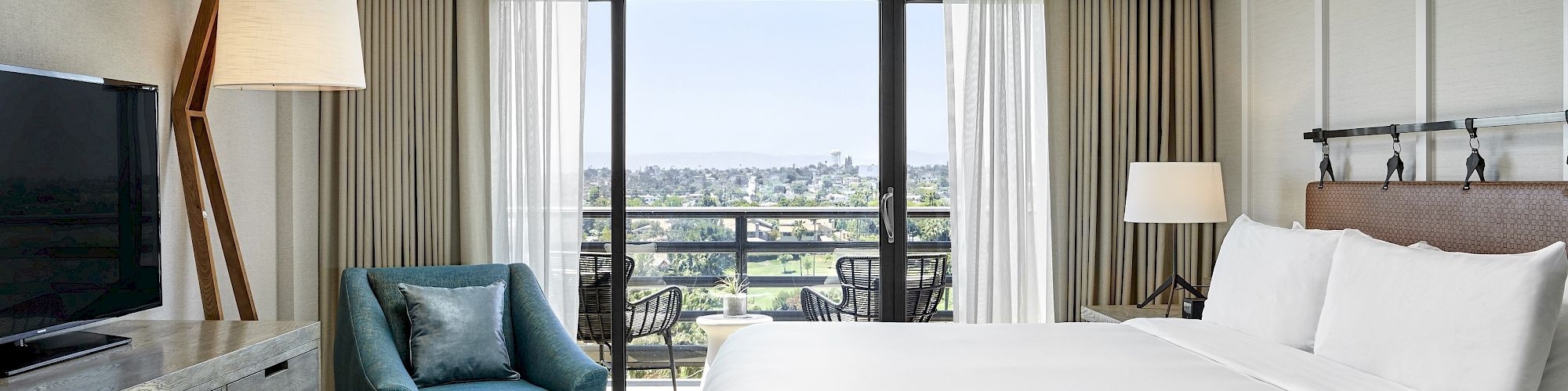 A modern hotel room with a bed, a TV, a comfy chair, lamps, curtains, and a view of a balcony.