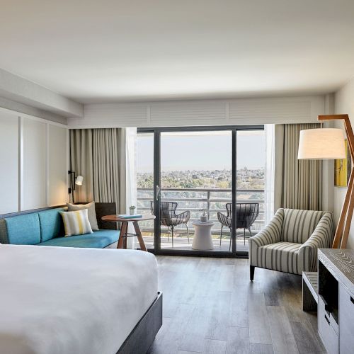 A modern hotel room with a bed, sofa, striped chair, large TV, and a balcony with a view. The decor is sleek and contemporary.