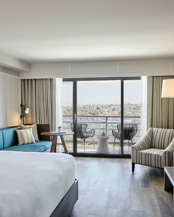 A modern hotel room with a bed, sofa, striped chair, large TV, and a balcony with a view. The decor is sleek and contemporary.