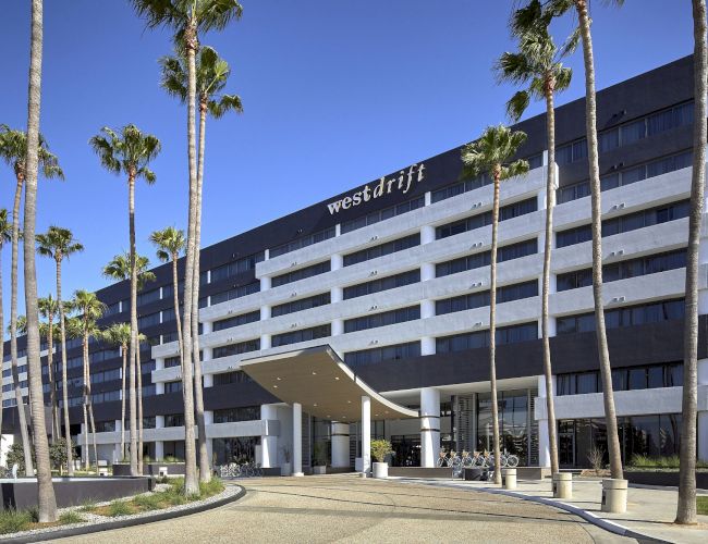 The image shows a modern multi-story hotel with palm trees lining the entrance under a clear blue sky.