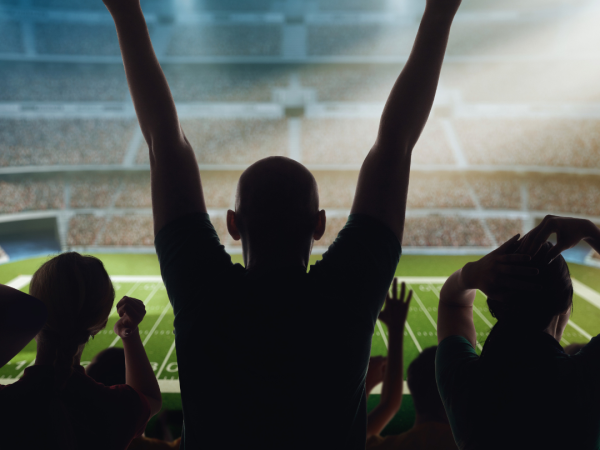 Fans in a stadium cheer, silhouetted against bright lights over a football field, creating an energetic atmosphere.