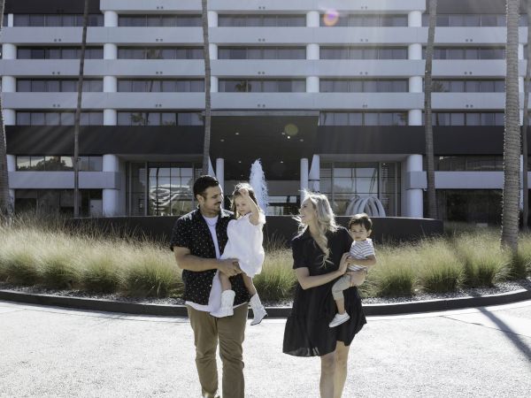 A family of four is walking together in front of a multi-story building with palm trees nearby and sunlight casting shadows.