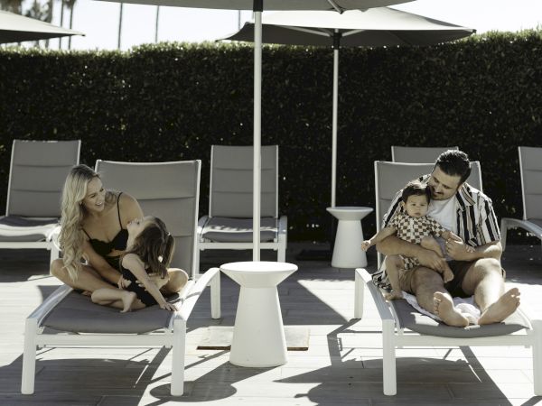 People are lounging on poolside chairs under umbrellas. A dog is sleeping on one side. Palm trees can be seen in the background.