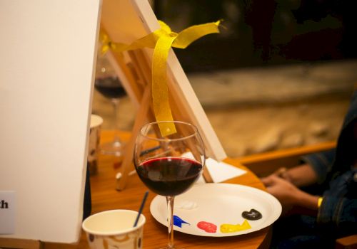 Blank canvases on easels, a palette with paint, a glass of wine, and a paper cup on a table suggest a painting session.
