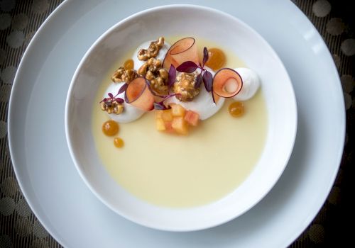 A plated creamy soup with delicate garnishes: sliced radish, flowers, almond slivers, micro herbs, and citrus droplets, served in a white bowl on a blue charger.